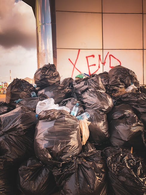 A large pile of black plastic rubbish bags filled with waste is stacked against a tiled wall outdoors. Several clear plastic bottles are visible protruding from the bags, indicating bottle recyclables