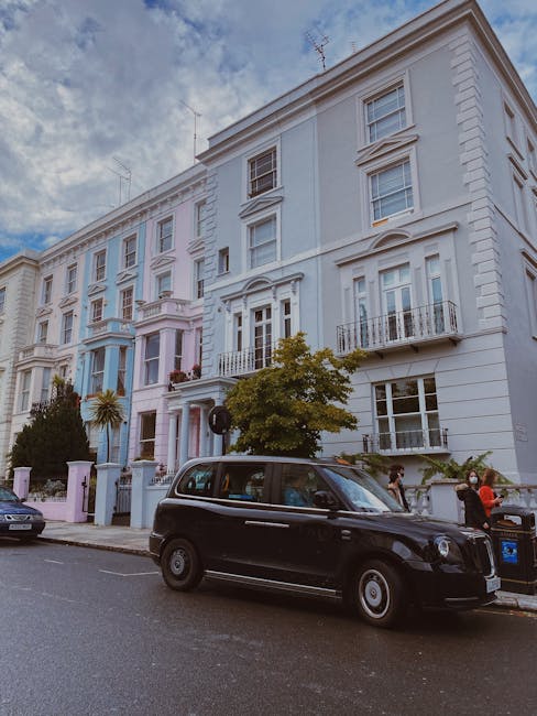 A busy street scene in an urban area with a row of multi-storey buildings lining both sides of the road, featuring a mix of shops, cafes, and businesses. The buildings are painted in various pastel an